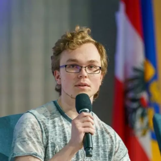 Tobias Winter spricht mit einem Mikrofon in der Hand bei einem Event. Im Hintergrund sind die österreichische und niederösterreichische Flagge zu sehen.